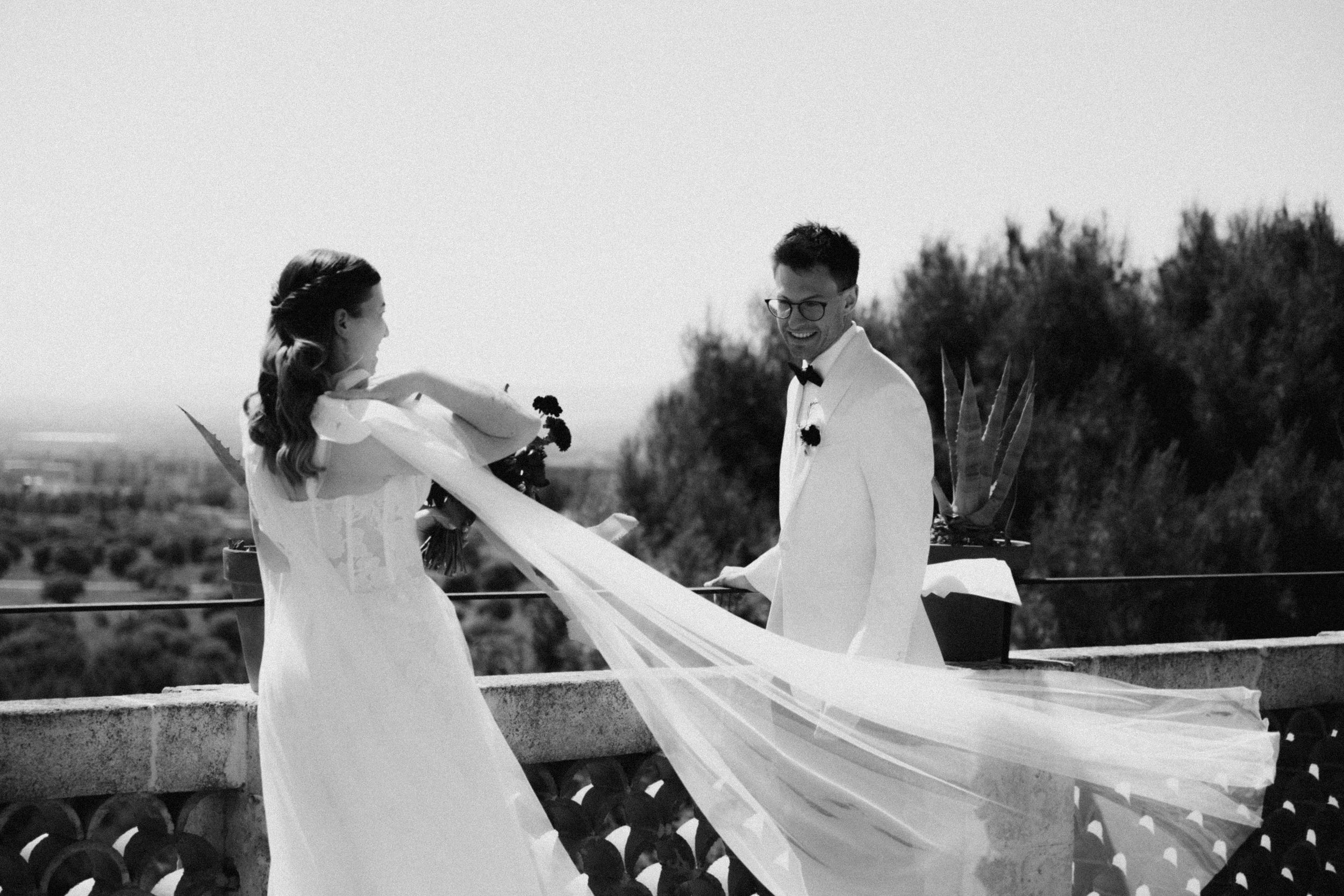 Black and white photo of bride and groom holding hands on a balcony with veil flowing in the wind.