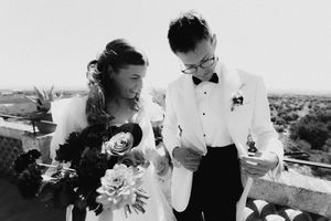 Black and white wedding photo of bride holding bouquet and groom in white tuxedo reading vows on outdoor balcony.