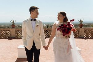 Bride and groom hold hands on a sunlit terrace, she holds red bouquet, he wears ivory tuxedo, scenic landscape behind.