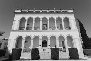 Black and white photo of a grand white colonial building with arched colonnade, decorative balustrade, and letter M signage.
