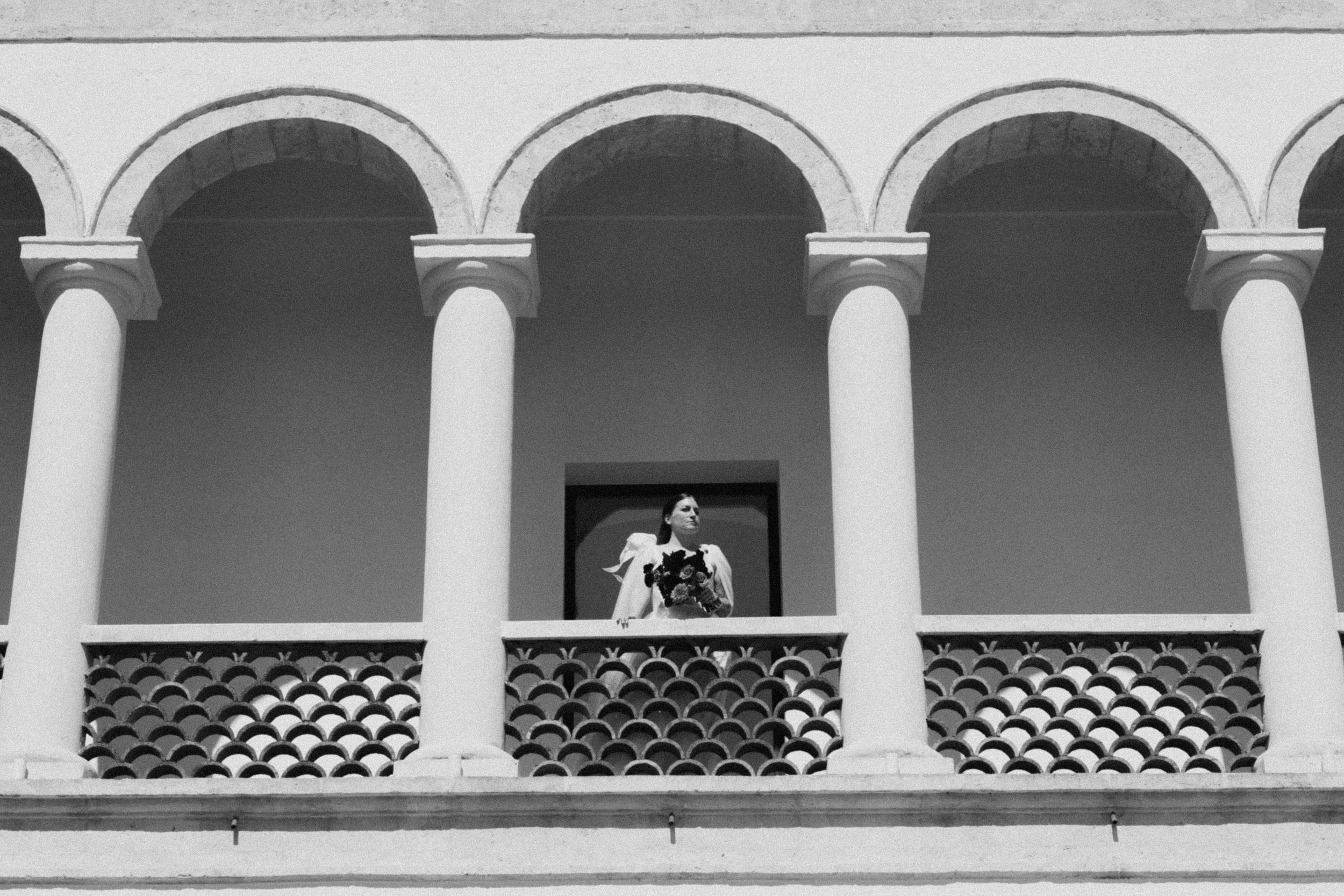Black and white photo of a person standing on an ornate balcony with arched columns of a classical building.