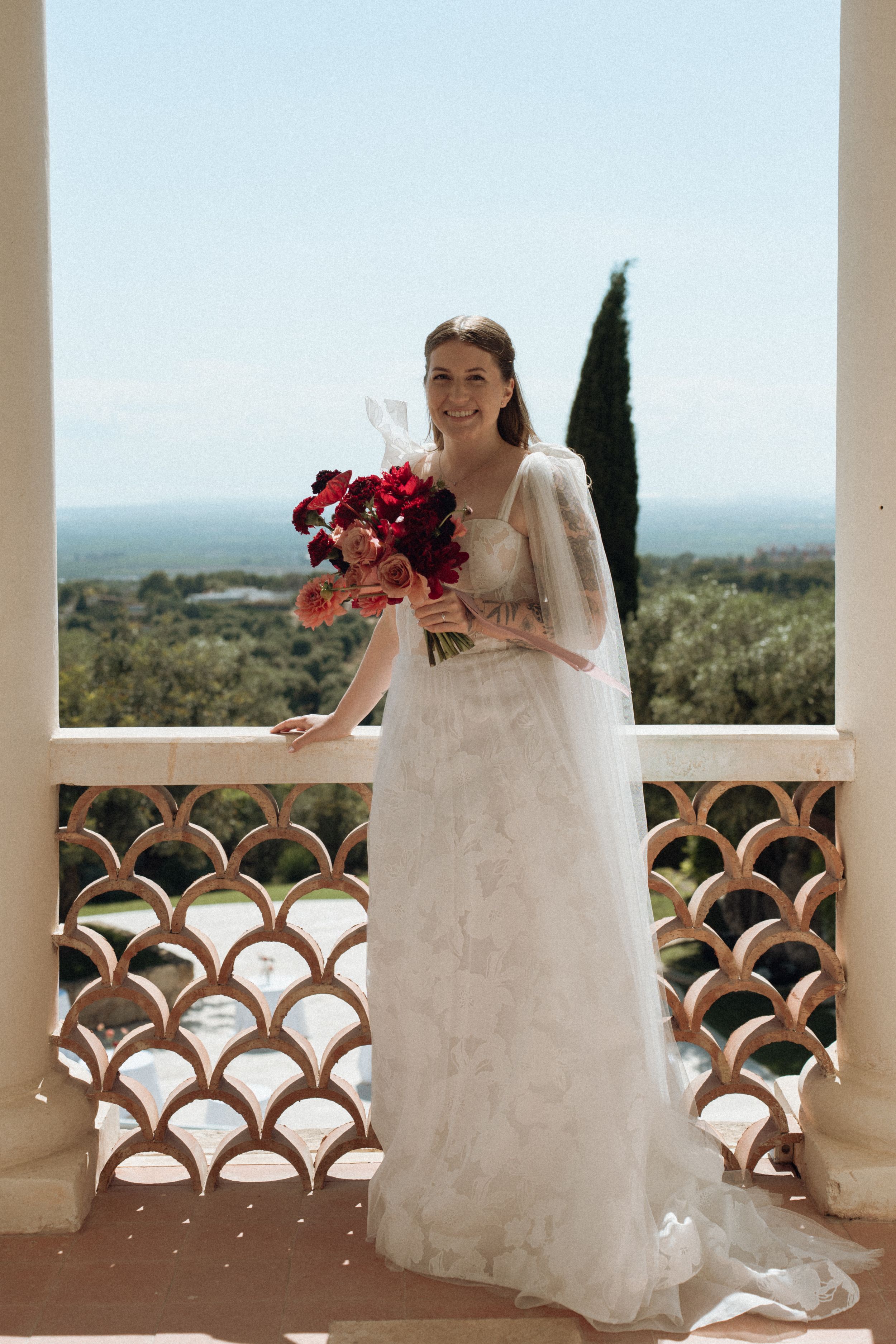 Bride in white gown holds red bouquet on ornate balcony with scenic landscape view on sunny wedding day.