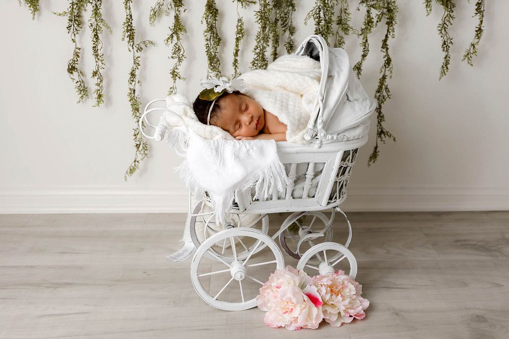 Newborn baby girl sleeping in white wicker pram with floral bouquet and hanging greenery backdrop.