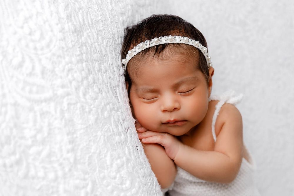 Sleeping newborn baby girl wearing a white headband wrapped in a soft white blanket.