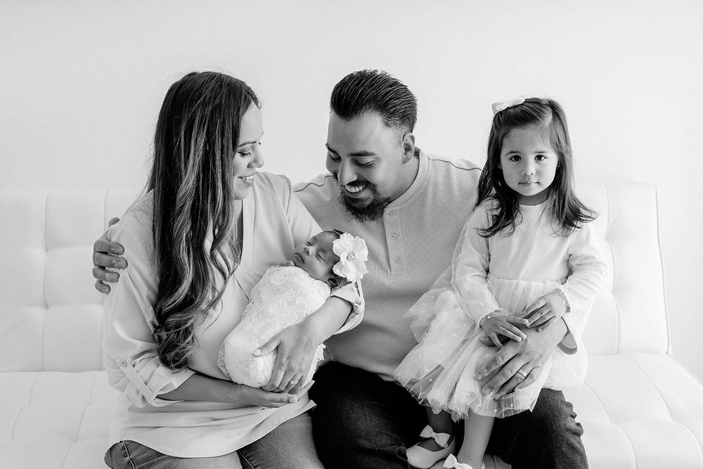 Black and white family portrait with parents and two young daughters, including a newborn baby, sitting together on a white couch.