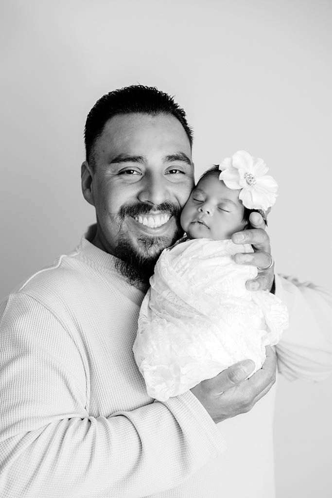 Smiling father holding sleeping newborn baby girl wrapped in white lace swaddle with floral headband, black and white photo.