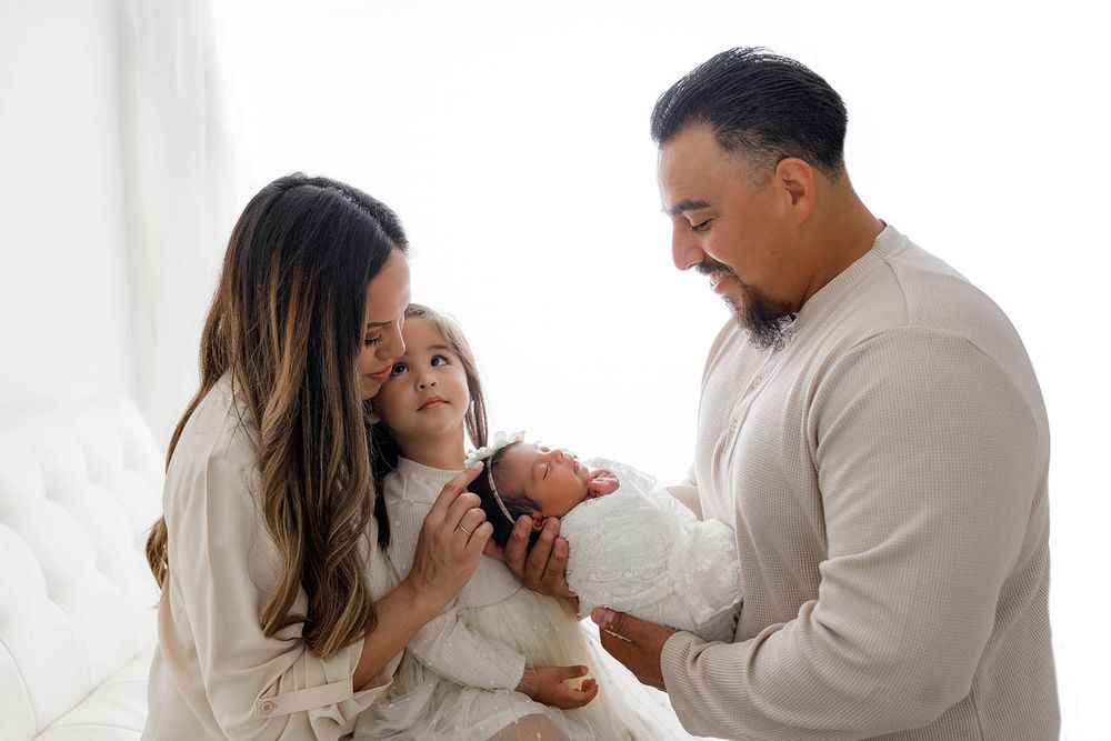 A happy family of four admires their newborn baby together in a bright white studio setting.