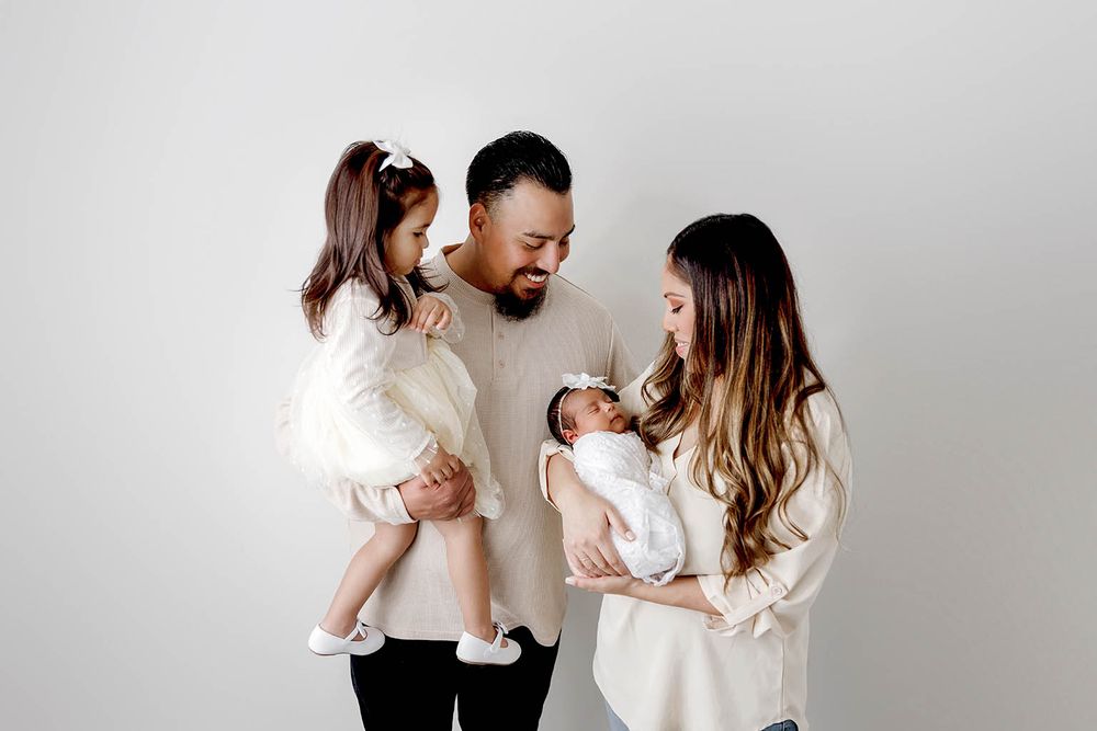 Happy family of four with toddler girl and newborn baby posing together in white studio setting.