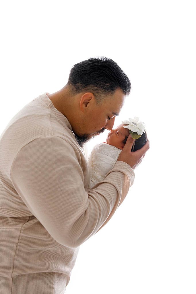 A father gently kisses his newborn baby wearing a white flower headband against a clean white background.