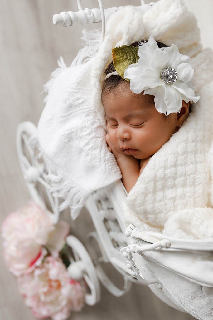 Sleeping newborn baby girl wrapped in white blanket wearing floral headband in decorative white pram.