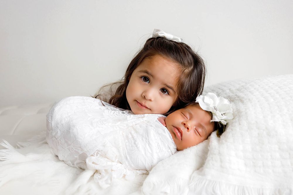 Young girl with dark hair gently resting beside sleeping newborn baby wrapped in white swaddle with floral headband.