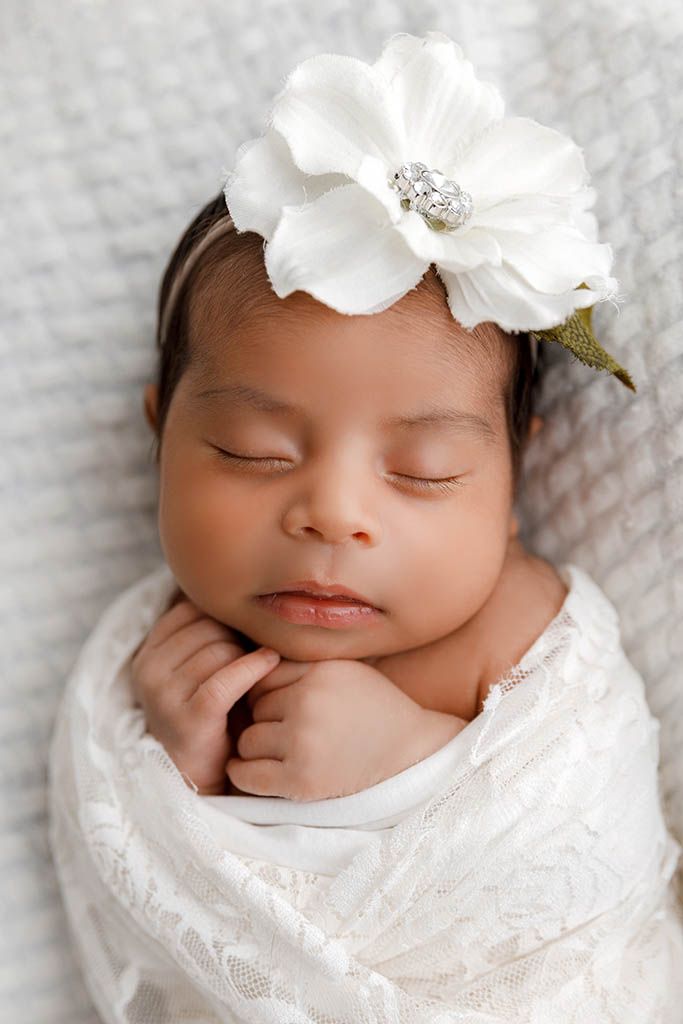 Peaceful sleeping newborn baby girl swaddled in white lace wrap wearing a large white flower headband with jewel.