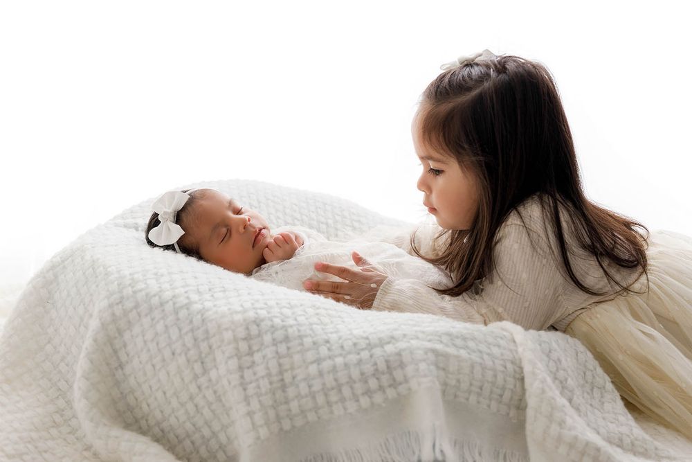 Young girl in white dress gazing lovingly at newborn sibling wrapped in white knit blanket.