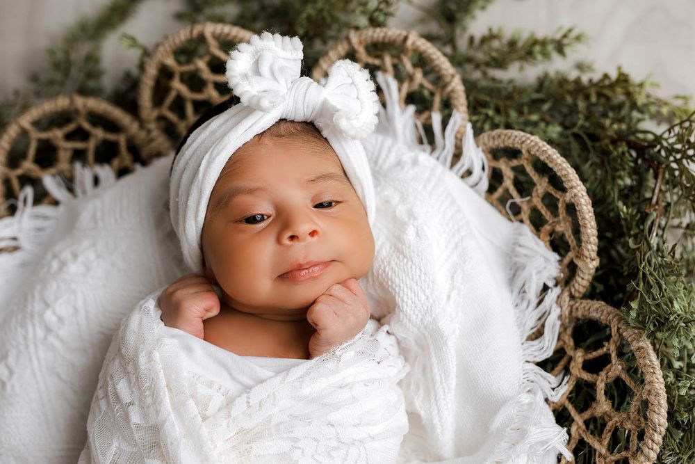 Alert newborn baby in white wrap and bow headband resting in wicker basket.