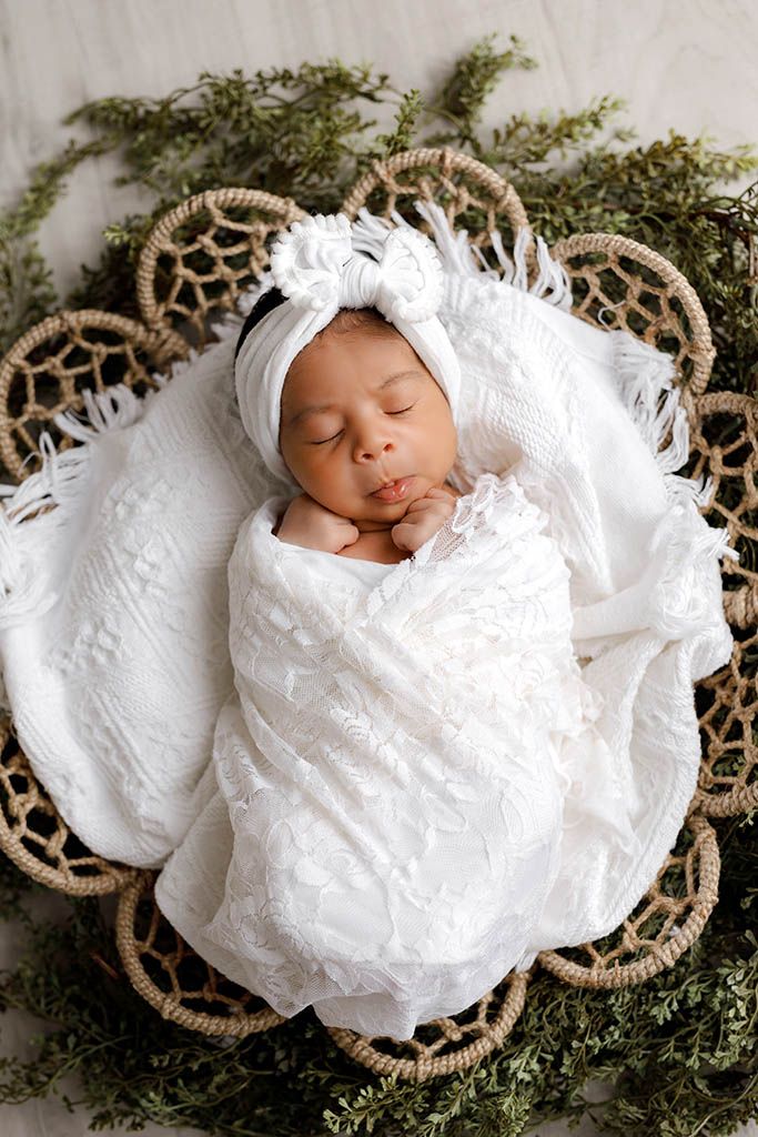 Sleeping newborn baby girl swaddled in white lace wrap with bow headband in woven basket.