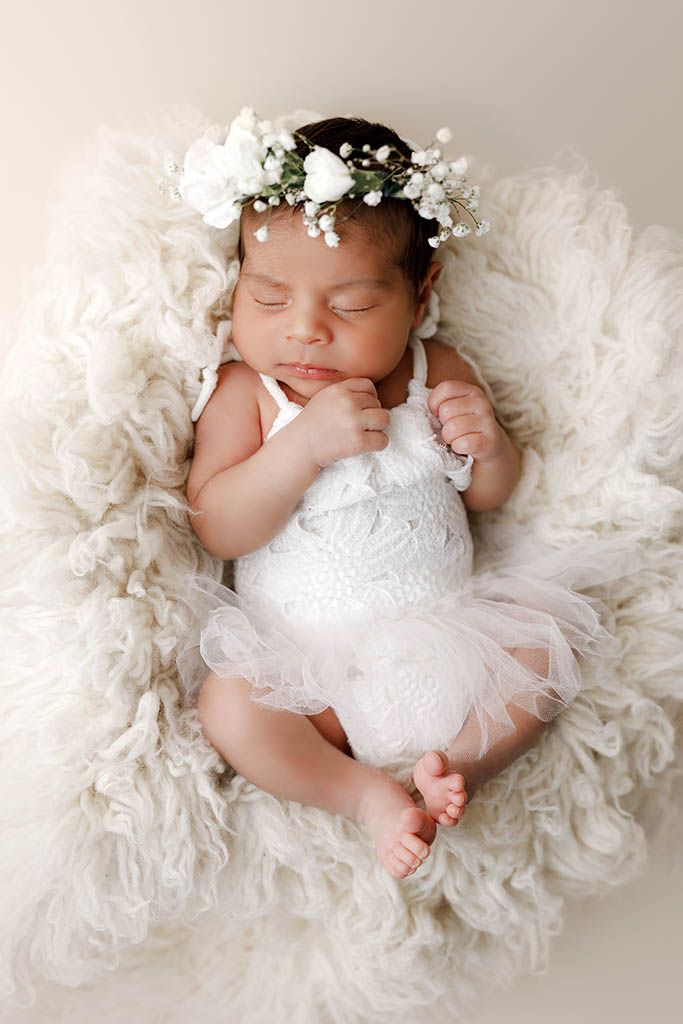 Sleeping newborn girl in white dress and floral crown on fluffy cream rug.