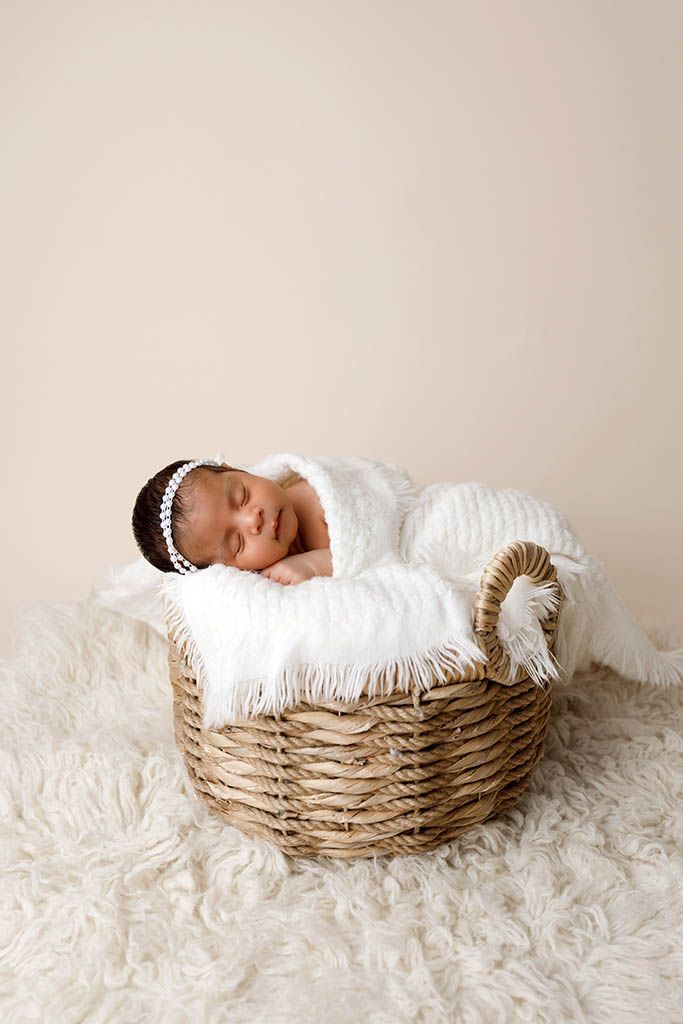 Sleeping newborn baby in white wrap resting in a wicker basket with fluffy white blanket on cream background.