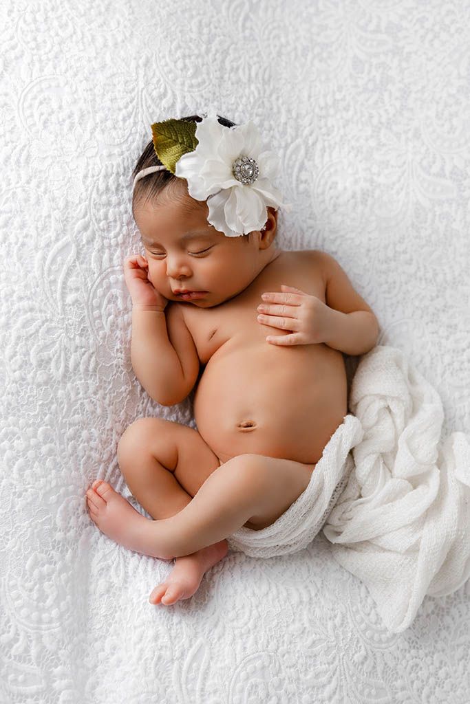 Sleeping newborn girl with white flower headband posed on white textured blanket.