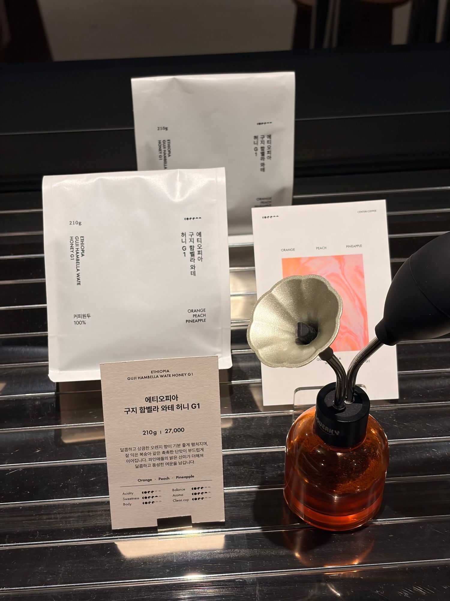 Korean coffee shop display with white packaging bags, a price card, and a decorative amber glass diffuser with funnel top.