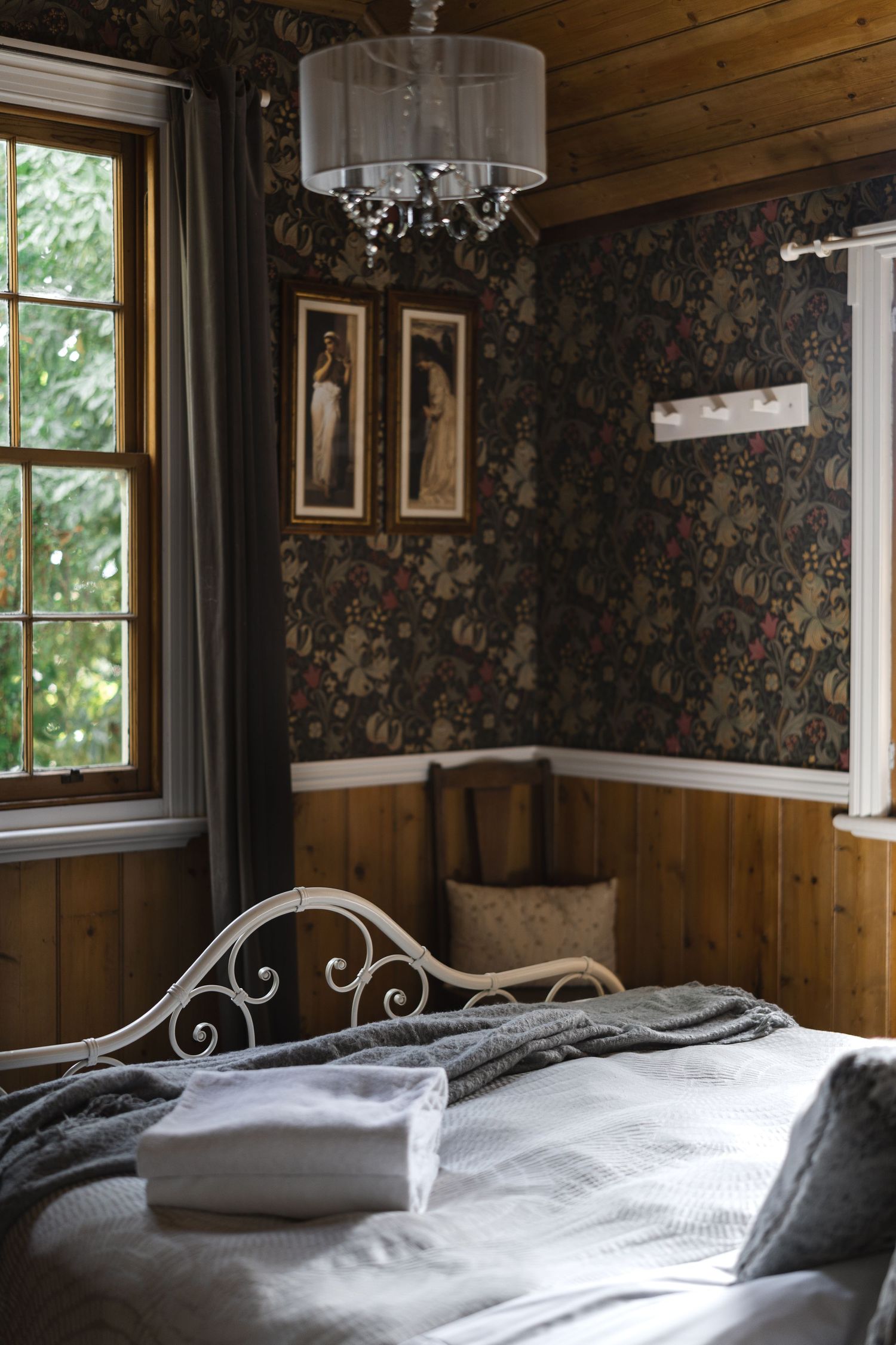 Elegant bedroom with dark floral wallpaper, ornate metal bed frame, chandelier, and wooden paneling.

Rosey Knoll