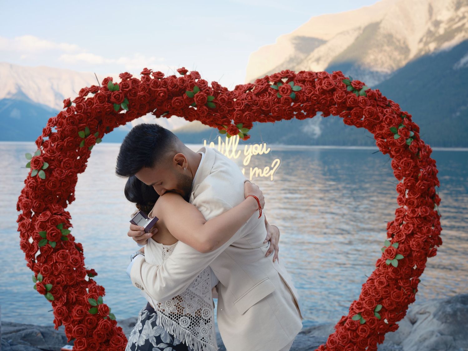 Couple embracing under a red rose heart arch with a Will You Marry Me sign, mountains and lake in background.
