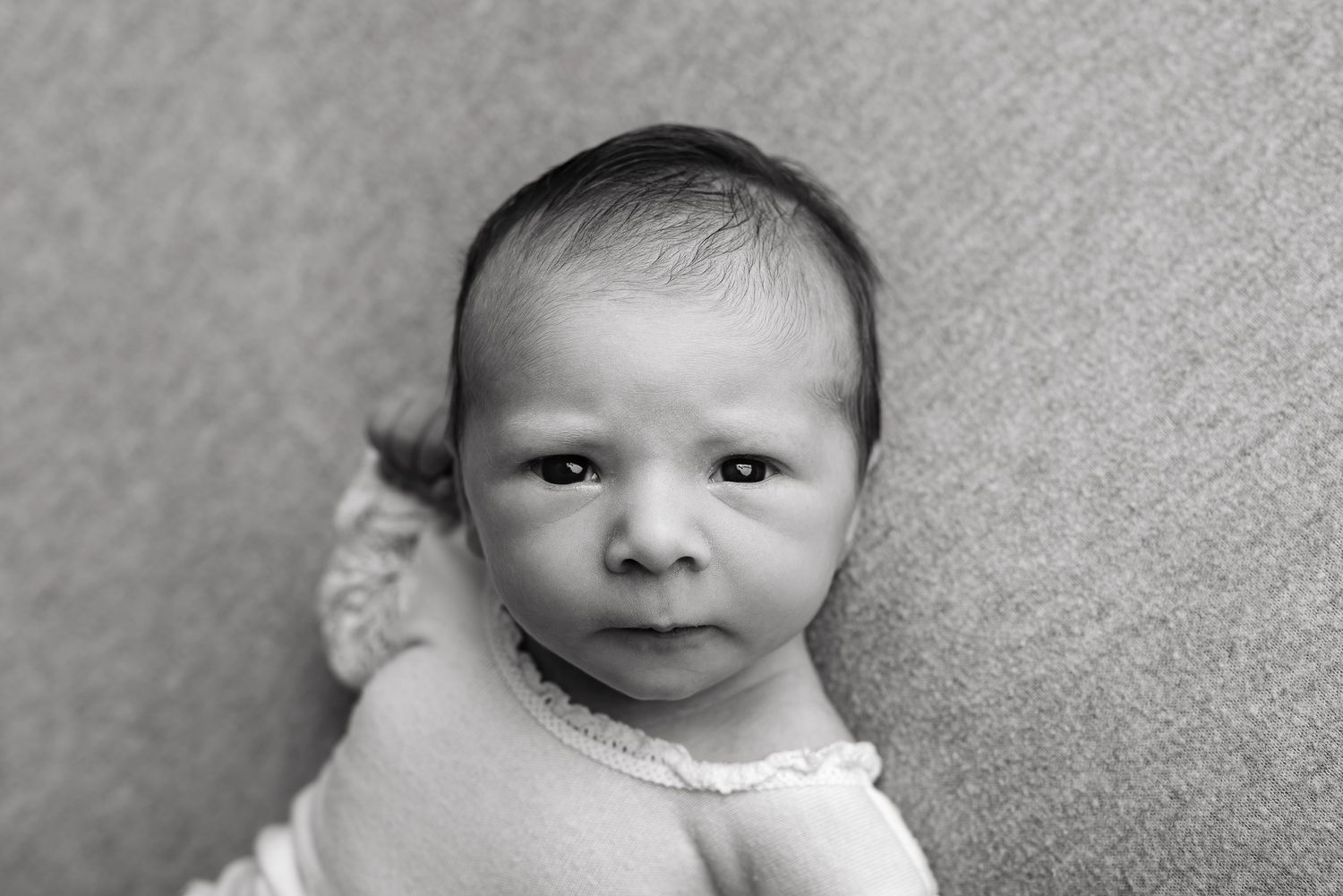 Black and white portrait of a newborn baby with dark hair, looking directly at the camera with a serious expression.