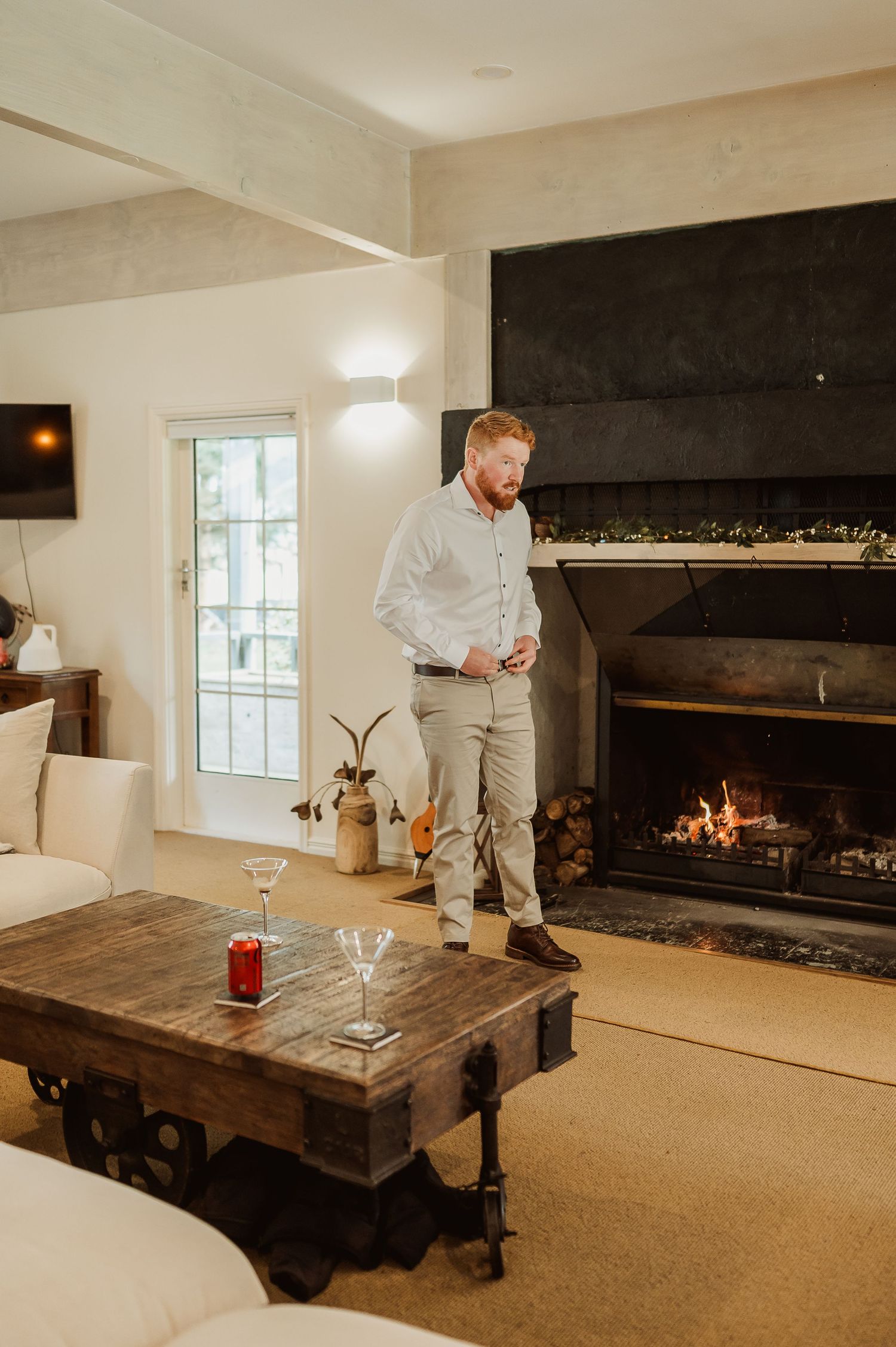 Man in white shirt stands by a lit fireplace in a cozy living room with a rustic coffee table and drinks.
