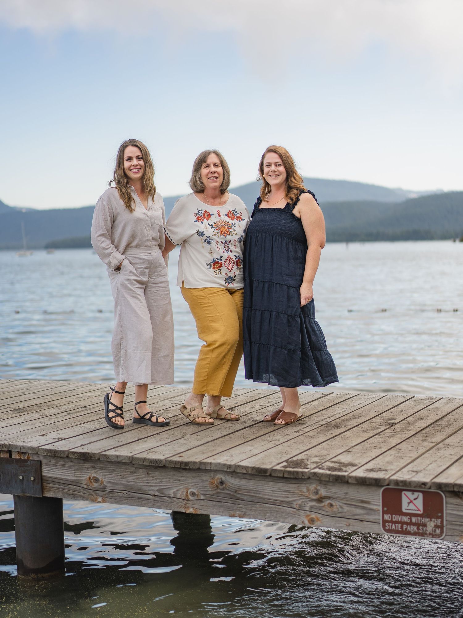 Three women smiling on a wooden dock by a lake with mountain views in the background.