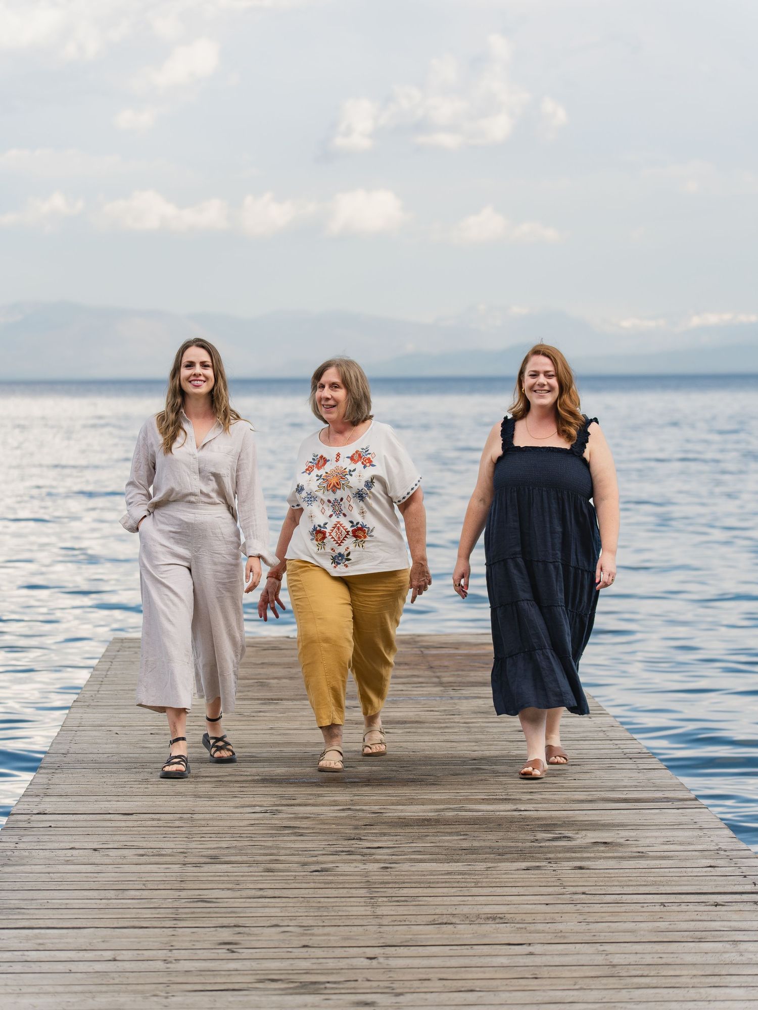 Three women walk confidently on a wooden dock over calm blue water, wearing stylish summer outfits.