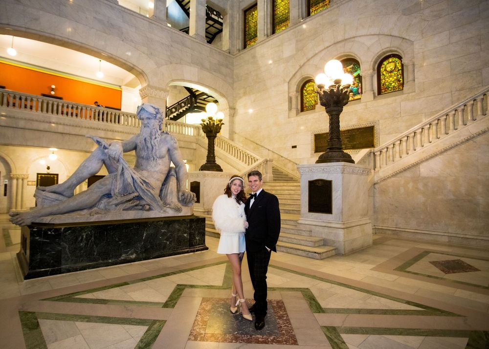 Couple posing in elegant marble rotunda with classical sculpture and grand staircase in ornate building interior.