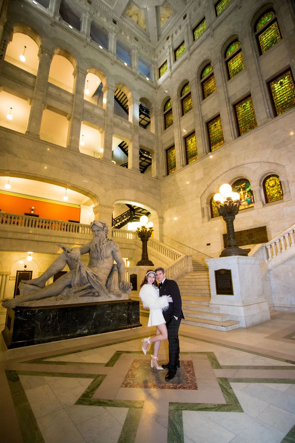 Bride and groom embracing in grand marble atrium with ornate stained glass windows and classical sculpture.