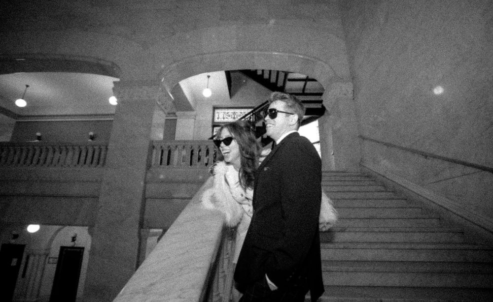 Black and white photo of a bride and groom in sunglasses walking up grand staircase at elegant wedding venue.