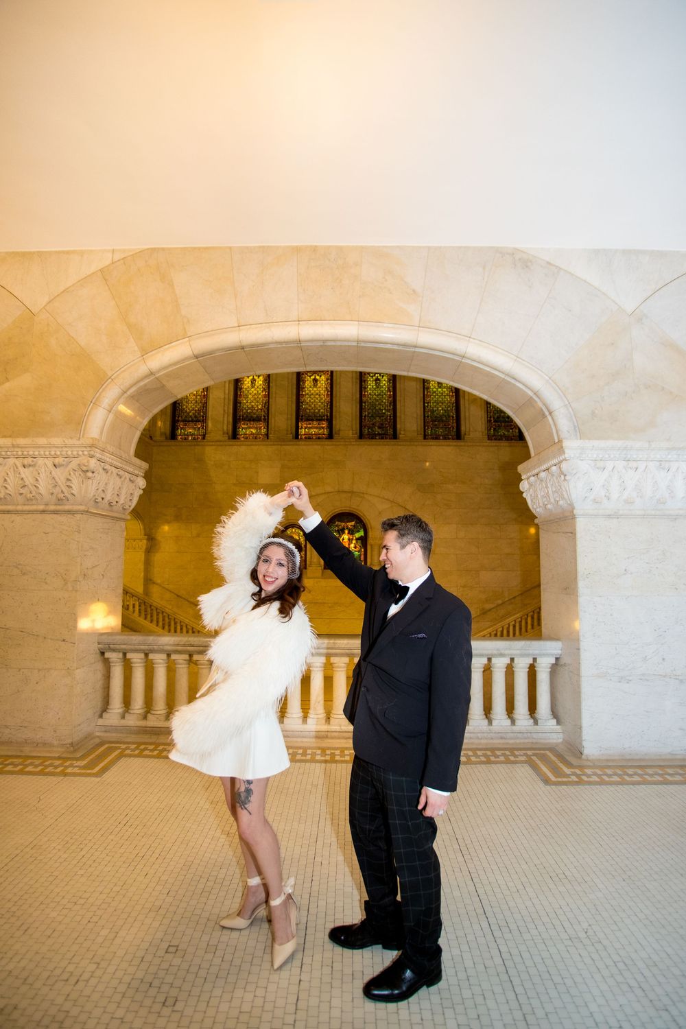 Couple dancing in elegant marble hall, bride in white mini dress with feather hat, groom in black tuxedo.