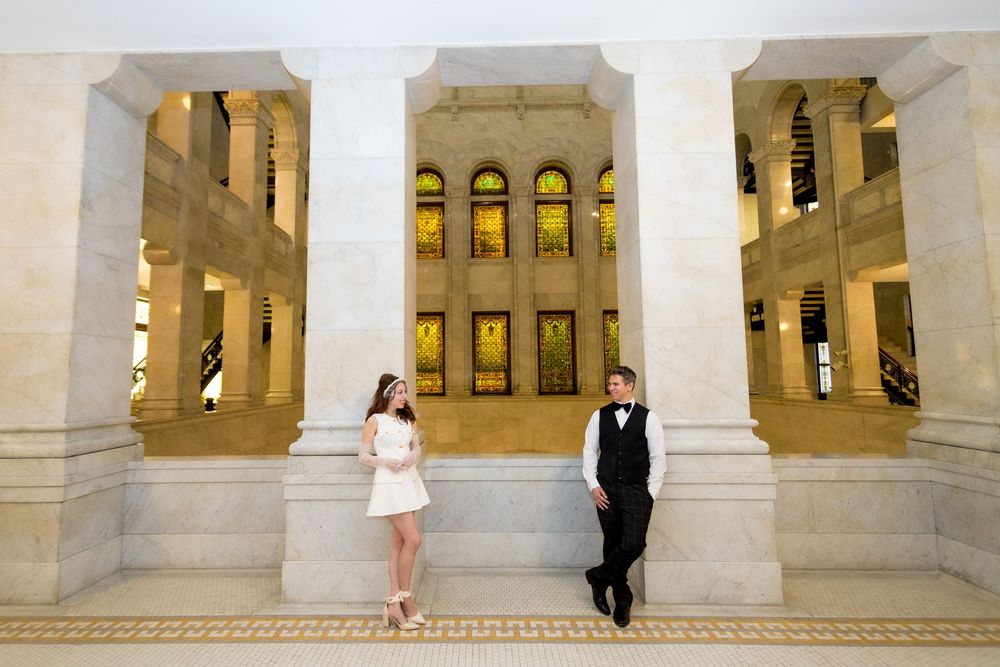 Couple poses in elegant marble hall with golden arched windows and tall columns for a wedding photo shoot.