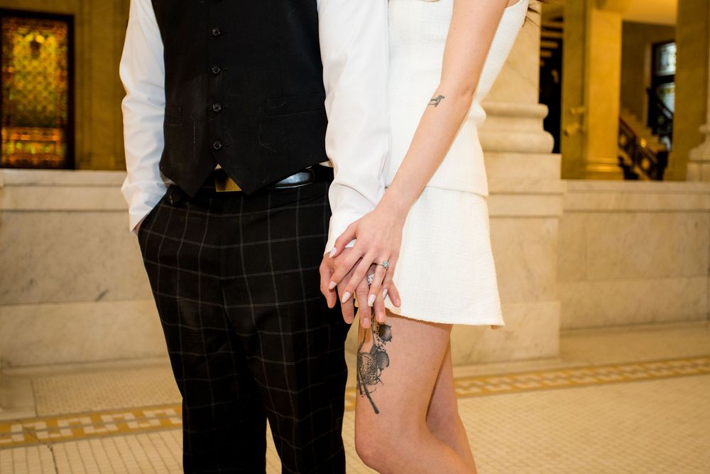 Couple holding hands, woman in white mini dress with rose tattoo, man in black vest and plaid pants.