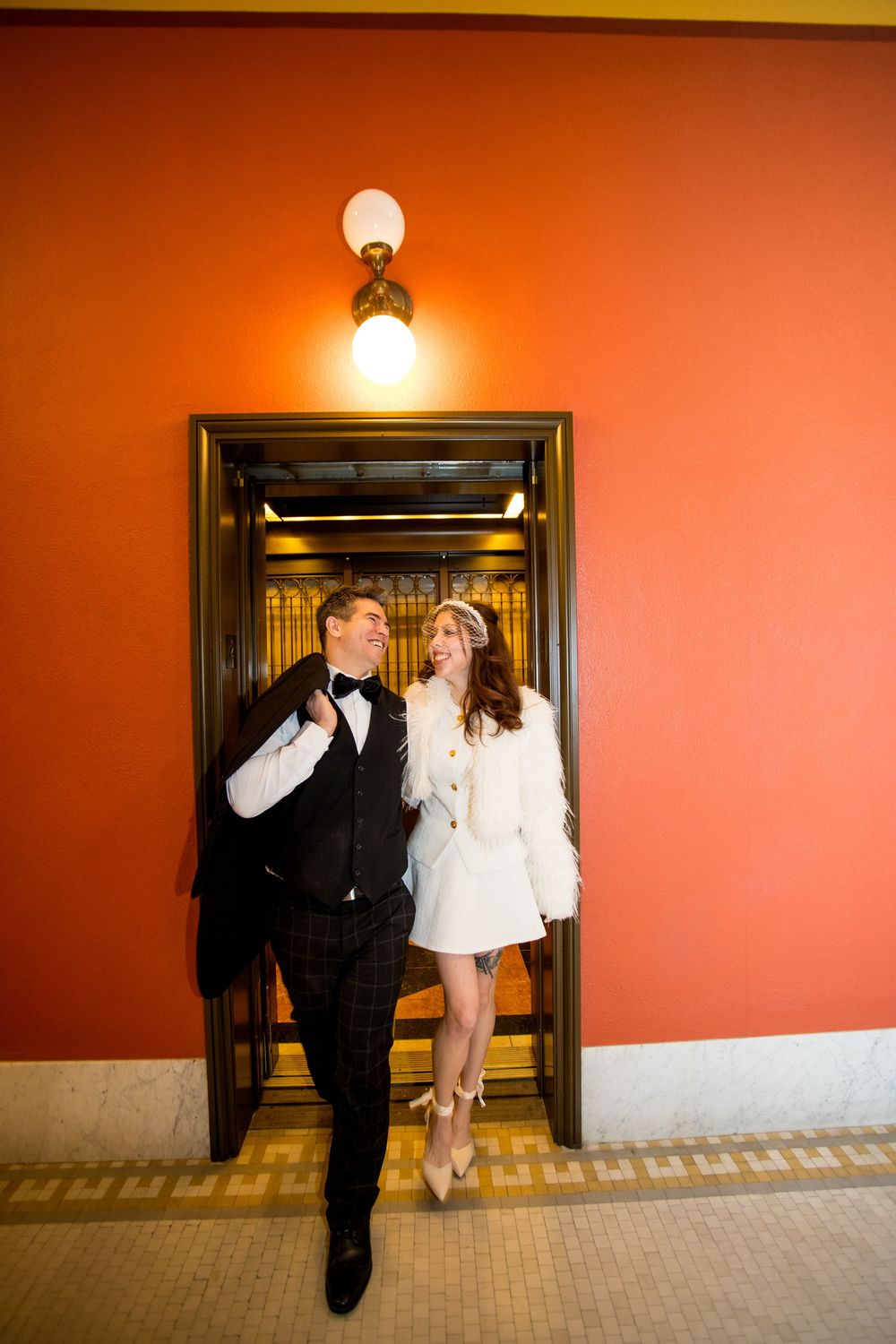 Bride and groom exit a vintage elevator, smiling together against a vibrant red wall.