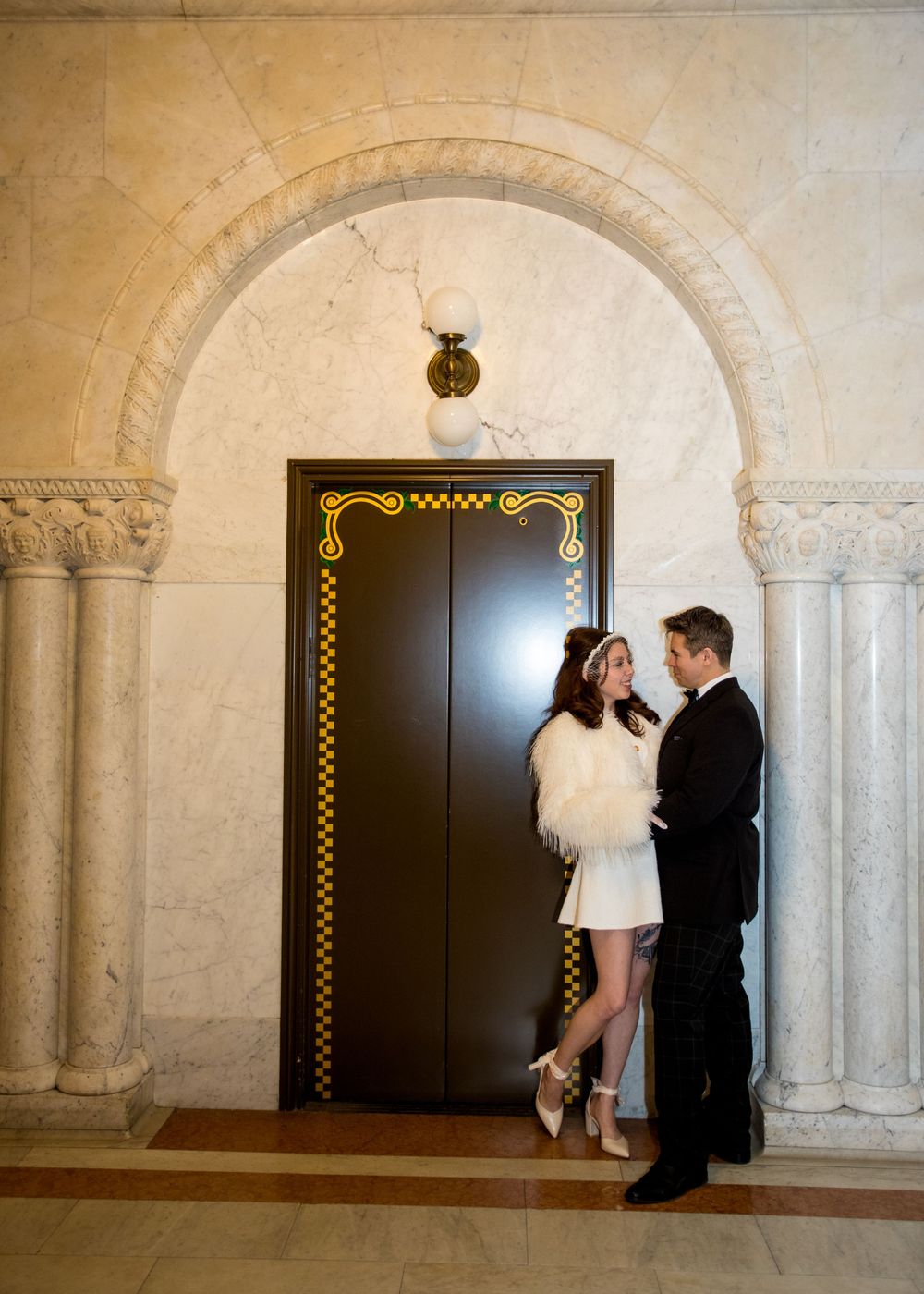 Elegant couple in formal attire posing by ornate elevator in marble-columned hall, bride in white mini dress with fur stole.