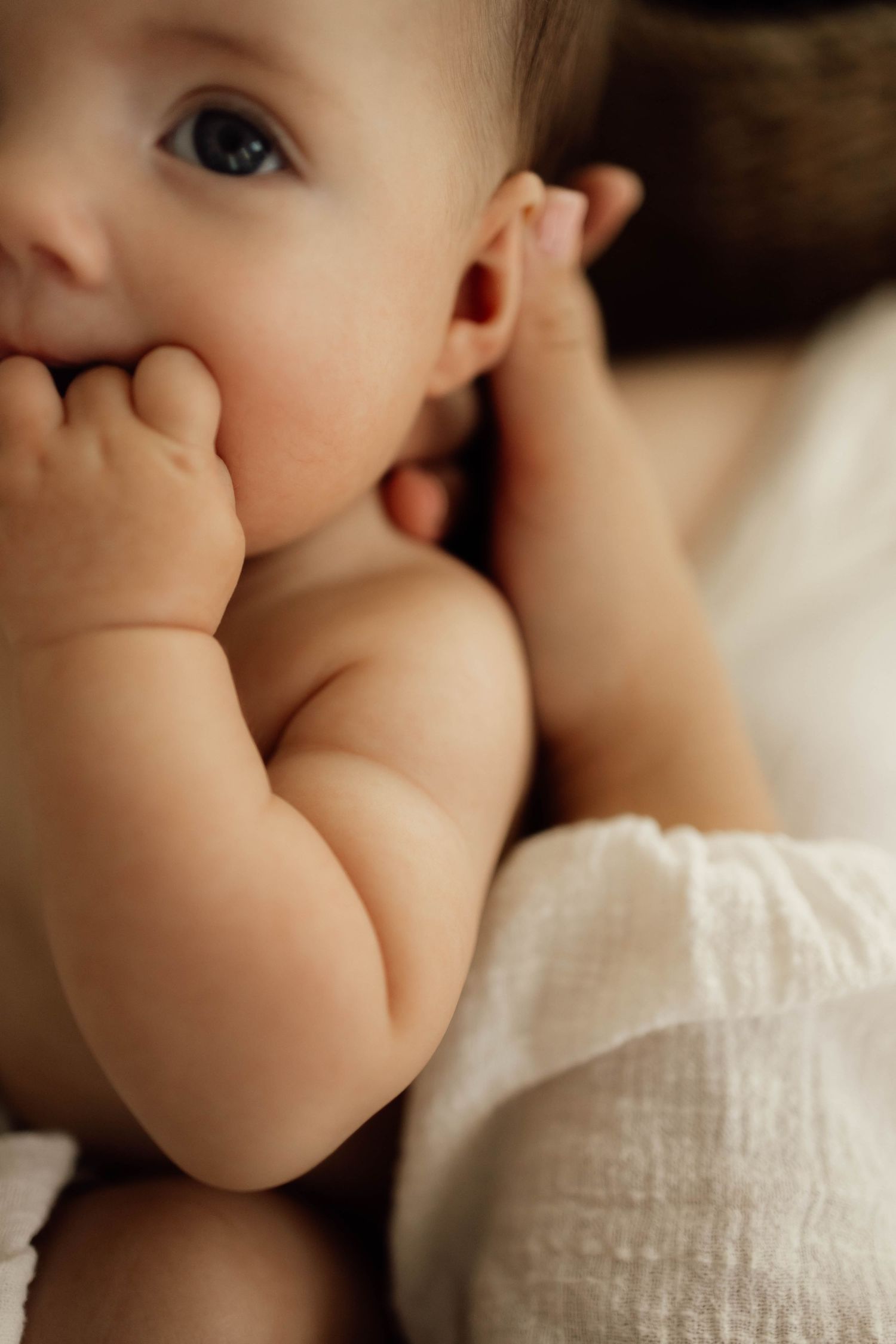 Adorable baby with blue eyes chewing on fingers, wearing white muslin diaper cover, soft warm lighting.