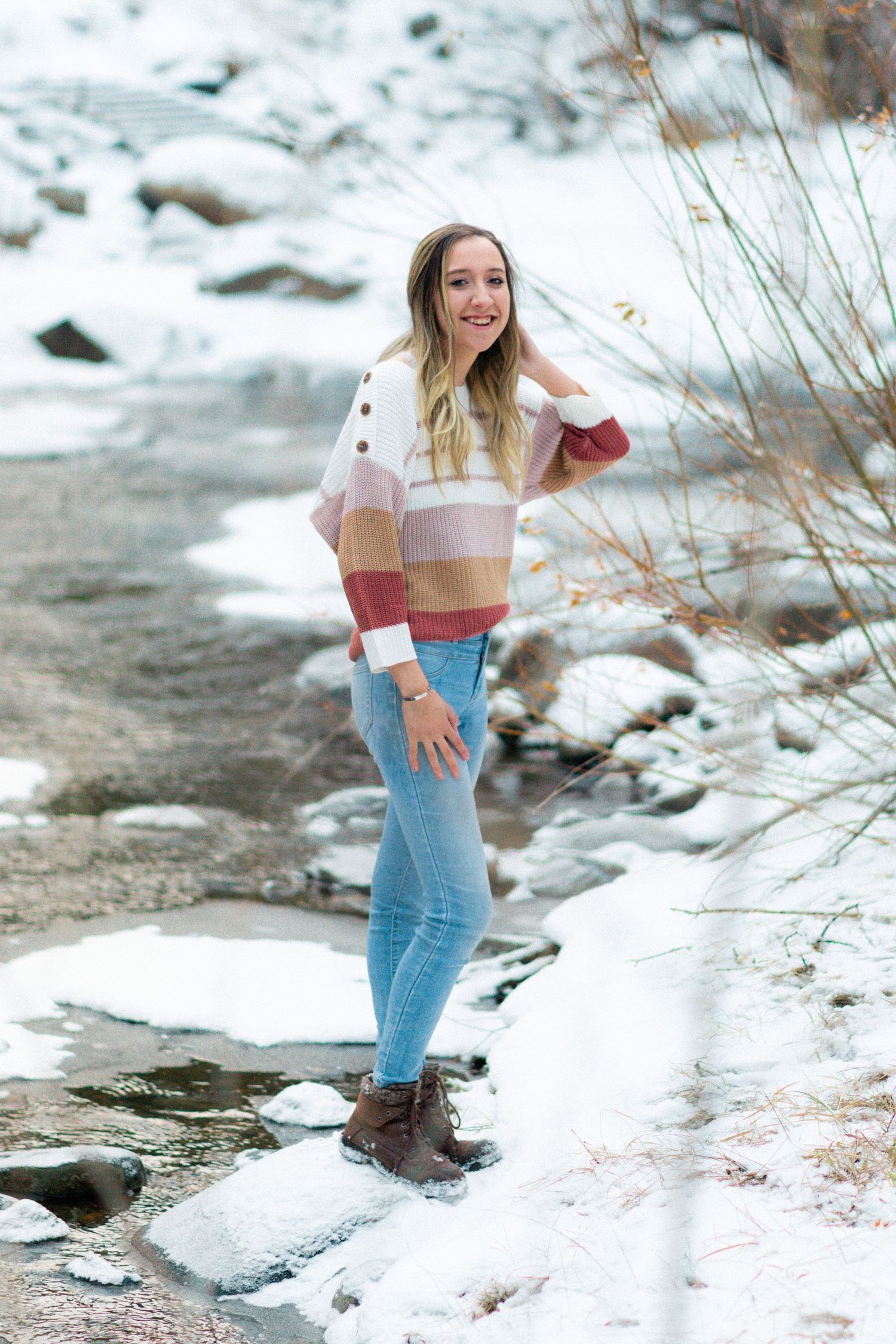 Young woman in striped sweater and jeans poses by snowy creek with rocks and bare branches in winter.