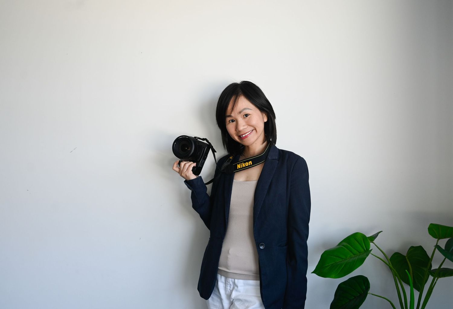 A smiling Asian woman in a navy blazer holds a Nikon camera, standing against a white wall near a green plant.
