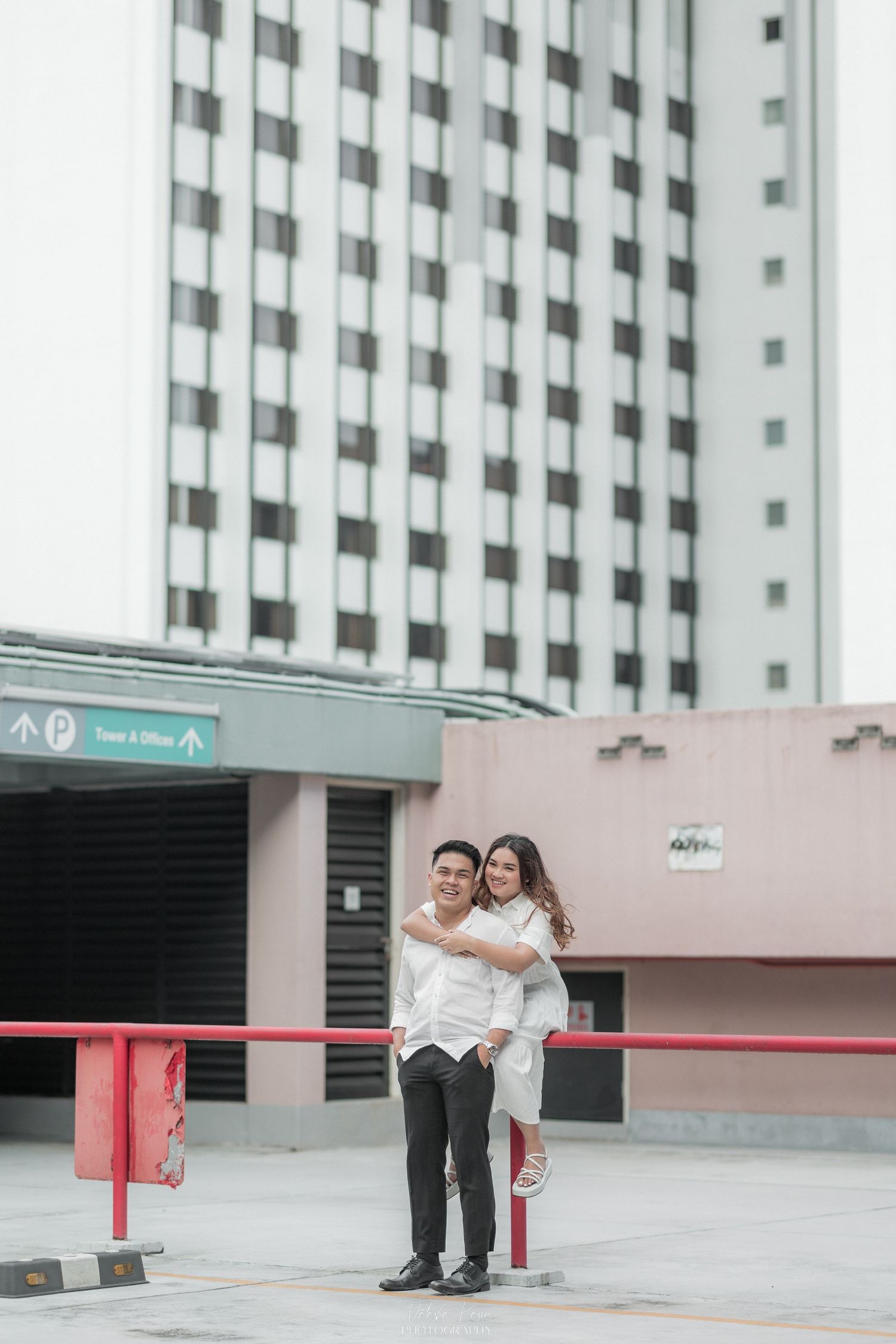 Couple in white outfits embracing in urban parking area with tall building backdrop.