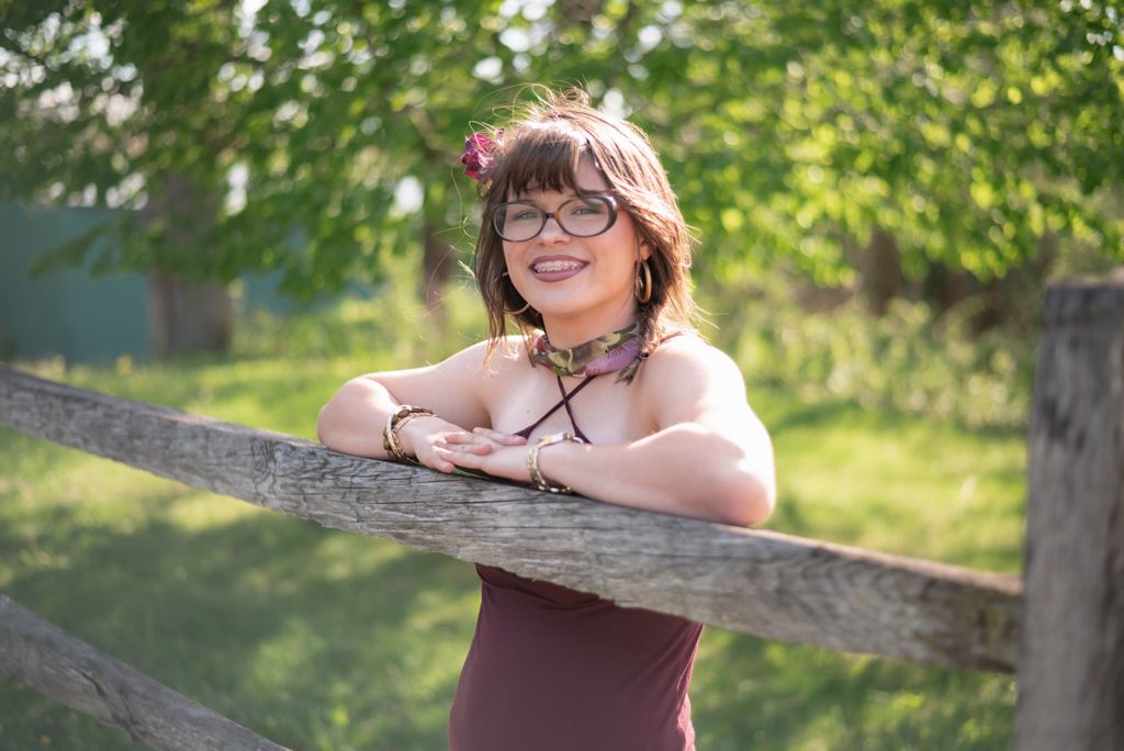 Young woman with glasses smiling outdoors, leaning on a wooden fence surrounded by lush green trees.