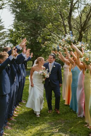 Wedding party cheers with raised bouquets as bride in white gown and groom in navy suit walk through floral arch.