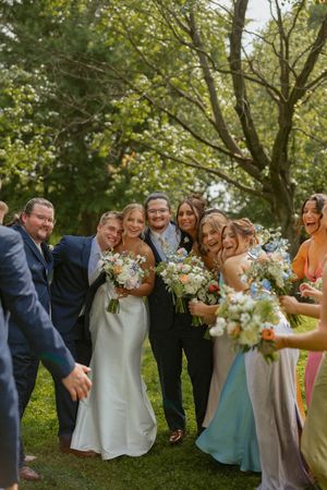 Joyful wedding party group photo outdoors with bride holding bouquet surrounded by bridesmaids and groomsmen.