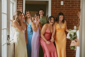 Bridesmaids in colorful satin dresses react with joy and excitement upon seeing the bride for the first time.