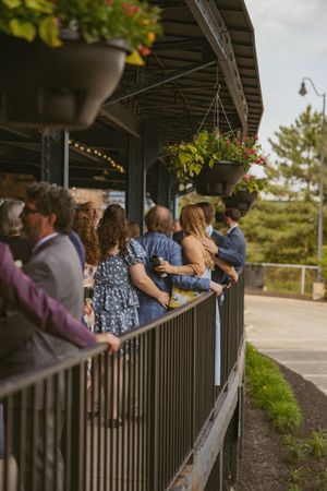 Wedding guests mingle on a decorated outdoor patio with hanging flower baskets and string lights above.