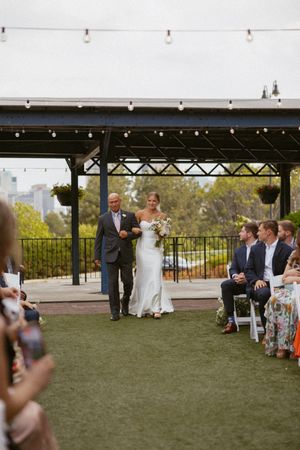Bride walking down outdoor aisle with escort at elegant rooftop wedding ceremony with string lights.