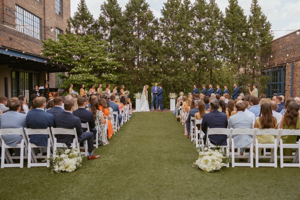 Outdoor wedding ceremony on green lawn with guests seated in white chairs, floral arrangements, and string lights at an urban venue.