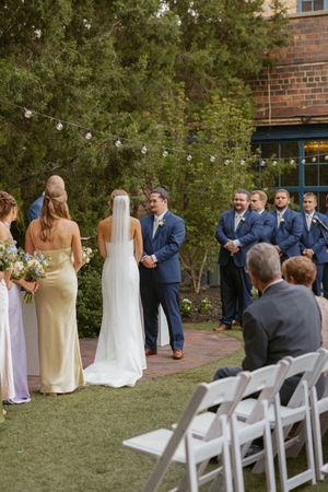 Bride in white gown and veil holds hands with groom in navy suit during outdoor wedding ceremony on brick path.