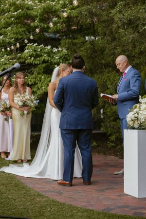 Bride and groom exchange vows outdoors with officiant reading from book, bridesmaids in pastel dresses nearby.